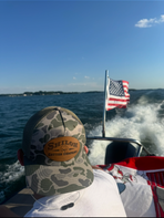Person on a boat with an American flag, wearing a camouflage cap, against a blue sky and water background.